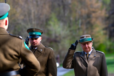 Dwie osoby w mundurze Straży Granicznej oddaje honor. Z lewej trony tyłem stoi mężczyzna w mundurze. W oddali widać drzewa.