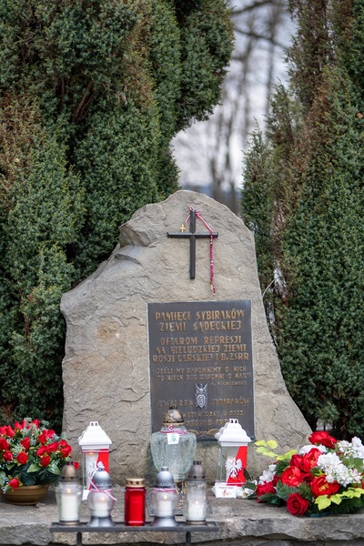 Na zdjęciu widać szary kamienny obelisk z czarną tablicą, na której umieszczone są złote litery. W tle widać krzewy w kolorze zielonym. Widać kolorowe kwiaty złożone pod obeliskiem oraz zapalone znicze.