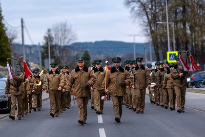 Na zdjęciu widać szeroką ulicę, po której idą ludzie ubrani w mundury, niektórzy niosą instrumenty. W tle widać drzewa, po prawej i lewej stronie zdjęcia widać fragmenty samochodów.
