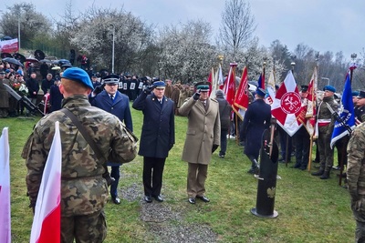 Na zdjęciu widać trzech mężczyzn w mundurach, którzy oddają honor. Po lewej stronie stoją ludzie, którzy trzymają w ręku sztandary, widać czarną tubę z przytwierdzonym na środku złotym orłem z rozpostartymi skrzydłami. W tle widać osoby, niektóre trzymają w ręku parasol. Widoczne są drzewa i zielona trawa. Po lewej stronie zdjęcia widać dwie flagi w kolorze biało-czerwonym.