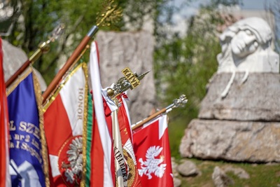 Na zdjęciu widać sztandary skierowane w prawą stronę. Po prawej stronie zdjęcia widoczny jest kamienny obelisk na którym umieszczona jest biała głowa nachylona w prawą stronę. Widać zieloną trawę i zielone drzewa.
