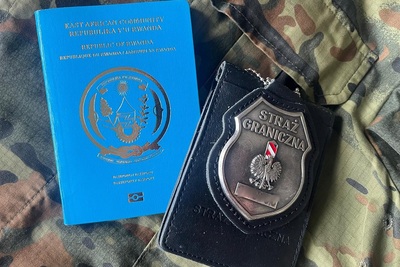 Na zdjęciu widoczny jest paszport koloru niebieskiego ze złotymi napisami oraz czarna legitymacja z metalowym emblematem. Przedmioty leżą na bluzie w kolorze moro.