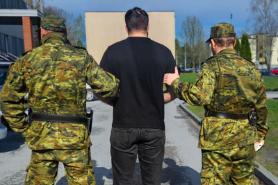 Dwóch funkcjonariuszy SG ubranych w mundur polowy prowadzi zatrzymanego przez podjazd do pomieszczeń SG, w tle widać budynki oraz samochody stojące na parkingu