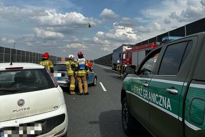 na zdjęciu widoczne jest zdarzenie drogowe. wypadek ma miejsce na autostradzie. na pierwszym planie widoczny pojazd służbowy straży granicznej z napisałem straż graniczna. w tle widoczne są samochody biorące udział w kolizji i helikopter ratownictwa medycznego który podchodzi do lądowania.