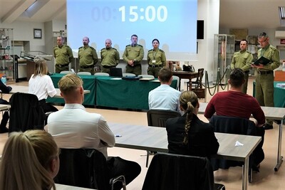na pierwszym planie widoczni są kandydaci aplikujący do służby w straży granicznej. na drugim planie widoczna jest komisja egzaminacyjna  złożona z funkcjonariuszy straży granicznej. do kandydatów mówi przewodniczący komisji egzaminacyjnej. całość odbywa się w sali odpraw karpackiego oddziału straży granicznej. na telebimie widoczny jest czas do zakończenia testu.