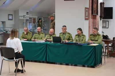 na zdjęciu widoczni są funkcjonariusze straży granicznej wchodzący w skład komisji egzaminacyjnej, którzy siedzą przy stole. na pierwszym planie widoczna jest kandydatka ubiegająca się do służby w straży granicznej, która siedzi przed komisją i odpowiada na ich pytania. całość odbywa się w sali odpraw Karpackiego Oddziału Straży Granicznej.