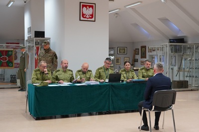 na zdjęciu widoczni są funkcjonariusze straży granicznej wchodzący w skład komisji egzaminacyjnej, którzy siedzą przy stole. na pierwszym planie widoczny jest kandydat ubiegający się do służby w straży granicznej, który siedzi przed komisją i odpowiada na ich pytania. całość odbywa się w sali odpraw Karpackiego Oddziału Straży Granicznej.