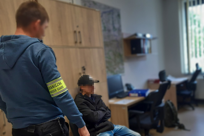 Na pierwszym planie zdjęcia jest funkcjonariusz straży granicznej ubrany w niebieską bluzę na prawej ręce ma założoną żółtą odblaskową opaskę z napisem straż graniczna polish border guard, na drugim planie siedzi na krześle  zatrzymany ubrany w czarną  bluzę oraz czarna czapkę w tle widać pomieszczenia służbowe straży granicznej