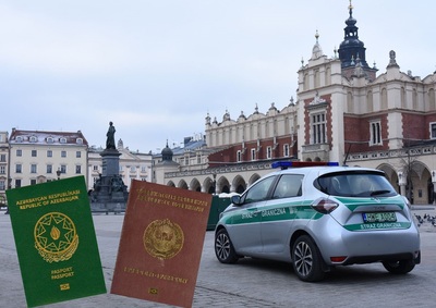 Na pierwszym planie widać paszporty koloru zielonego i bordowego z napisami koloru złotego na drugim planie samochód straży granicznej koloru srebrnego z zielonymi wstawkami i napisem straż graniczna w tle widać budynki krakowskiego starego miasta oraz sukiennice krakowskie
