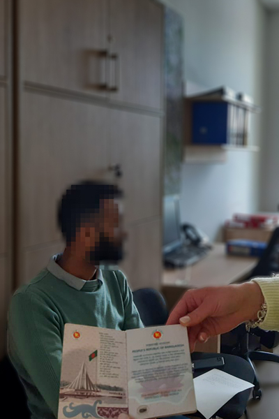 Na pierwszym planie Kobieca dłoń trzyma dokument paszportowy, na drugim planie zatrzymany cudzoziemiec siedzi na krześle zdjęcie wykonane w pomieszczeniach straży granicznej.