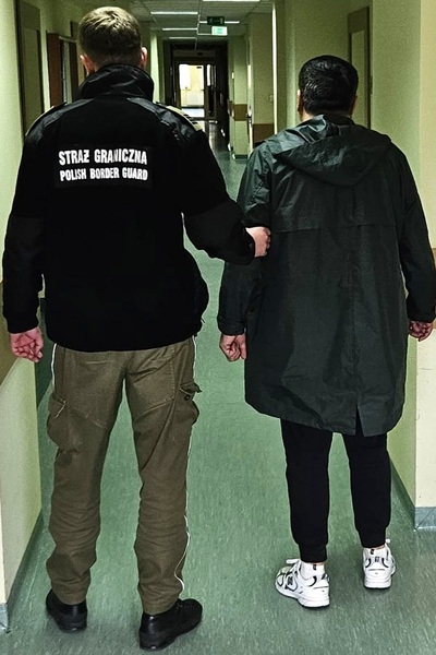 Funkcjonariusz ubrany w polar koloru czarnego z białymi napisami straż graniczne polish border guard, oraz spodnie koloru khaki, funkcjonariusz prowadzi pod rękę zatrzymanego cudzoziemca ubranego w ciemne ubranie, osoby znajdują się w pomieszczeniach straży granicznej