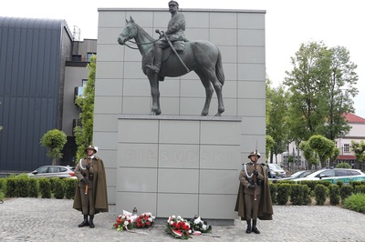 na zdjęciu widoczni są funkcjonariusze straży granicznej  tworzący posterunek honorowy. stoją na baczność. ubrani są w mundur galowy wraz z peleryną oraz kapeluszem. w dłoniach trzymają karabin reprezentacyjny. za nimi widoczny jest pomnik marszałka Piłsudskiego przedstawia marszałka który siedzi na koniu. w tle widać wielki mur który tworzy tło dla pomnika.