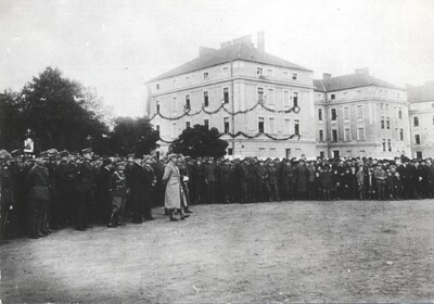 zdjęcie ukazuje fotografie z czasów między wojennych na której widoczny jest marszałek Piłsudski który wizytował koszary w nowym Sączu. w tle widoczny jest tłum ludzi oraz budynek koszar.