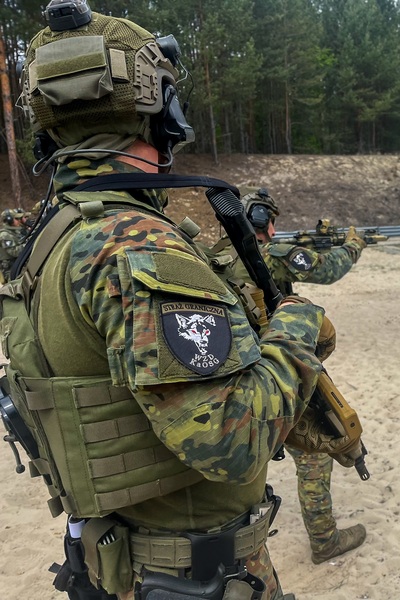 stojący funkcjonariusz straży granicznej ubrany w umundurowanie specjalistyczne  z bronią długą eksponuje ramię na którym jest naszywka z napisem straż graniczne  WZd Nowy Sącz