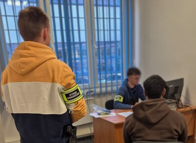 Jedne  funkcjonariusz w cywilnym ubraniu przesłuchuje zatrzymanego hindusa, siedzącego przy biurku. Drugi funkcjonariusz w cywilnych ubraniach stoi za cudzoziemcem.