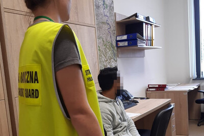 Pomieszczenia biurowe Placówki sg w tarnowie. Zatrzymany cudzoziemiec siedzi na krześle. pilnuje go funkcjonariuszka w kamizelce odblaskowej.