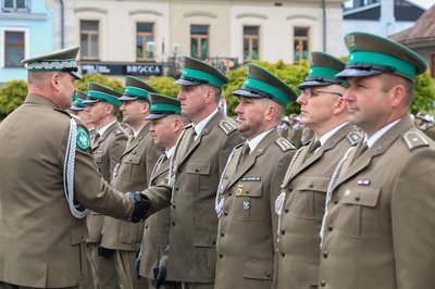 Na zdjęciu widać dziewięć osób, które ubrane są w mundury. Dwaj mężczyźni podają sobie ręce, na których mają czarne rękawiczki. W tle stoi rząd budynków. Widoczne są zielone drzewa.