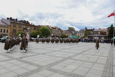 Na zdjęciu widać grupę osób ubraną w mundury, w rękach trzymają  instrumenty. W tle widać budynki i zielone drzewa. Z prawej strony zdjęcia widać flagę biało-czerwoną.