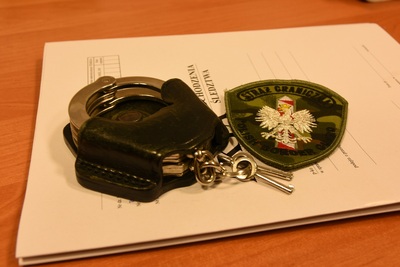 Na blacie biurka leżą akta sprawy na których znajdują się kajdanki, oprawa legitymacji służbowej Straży Granicznej oraz emblemat SG