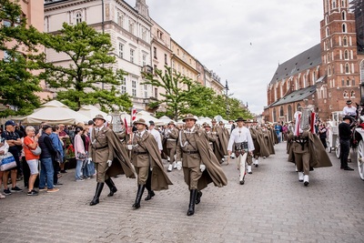 Orkiestra reprezentacyjna straży granicznej maszeruje i gra na instrumentach na rynku w Krakowie w tle kościół mariacki w koło ludzie robiący zdjęcia telefon
