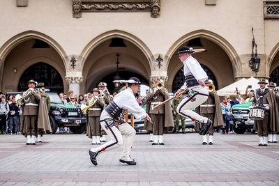 Dwóch funkcjonariuszy ubranych w strój góralski pokazujący wyszkolenie z ciupagami w tle orkiestra reprezentacyjna grająca podczas musztry paradnej