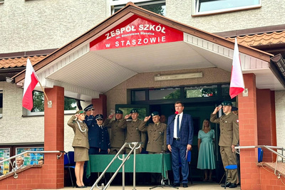 Oby stojące przed wejściem do szkoły oddają honor podczas grania hymnu narodowego, ubrani w mundury galowe różnych formacji oraz prowadzący uroczystość mężczyzna stojący na baczność ubrany w garnitur koloru granatowego, nad wejściem do szkoły widnieje napis z nazwą szkoły białe litery na czerwonym tle, na maszcie ściennym jest zawieszona flaga polski
