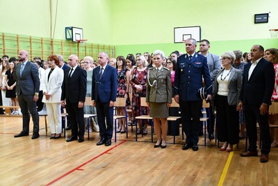 Osoby stojące na baczność podczas uroczystej zbiórki w hali sportowej, na pierwszym planie dwóch funkcjonariuszy straży granicznej i policji  ubrani w mundury galowe