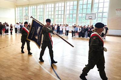 Poczet sztandarowy podczas przemarszu przez hale sportową ubrany w mundur polowy, w tle uczniowie szkoły