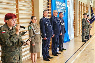 Osoby ubrane w mundury galowe podczas uroczystej zbiórki, wewnątrz hali sportowej osoba mówiąca przez mikrofon ubrana w mundur polowy(moro) w tle drabinki sportowe oraz napis rozpoczęcie roku szkolnego
