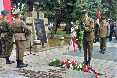 Żołnierz w zielonym berecie i mundurze składa wieniec z biało-czerwonych kwiatów pod tablicą pamiątkową umieszczoną na kamiennym pomniku. Obok stoją inni żołnierze w mundurach galowych, z karabinami na ramionach. Widać przygotowane już wcześniej wieńce ułożone u stóp monumentu. Całość odbywa się w spokojnej, poważnej atmosferze.