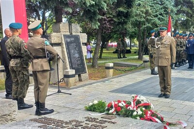 Wysoki oficer w mundurze galowym oddaje honory, salutując przed złożonymi kwiatami. W tle widoczna jest tablica pamiątkowa na kamiennym pomniku. Obok stoją żołnierze w zielonych beretach oraz oddział w szyku. Scena przedstawia moment oddania czci poległym przez gest wojskowego salutu.