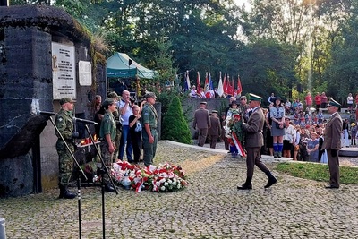 Uroczystość odbywa się przy betonowym schronie bojowym – zabytku z czasów II wojny światowej. Przy tablicach pamiątkowych umieszczonych na ścianach bunkra składane są wieńce i kwiaty. Obok stoją młodzi żołnierze oraz harcerze w mundurach. W tle widać poczty sztandarowe oraz licznie zgromadzoną publiczność, co świadczy o lokalnym i patriotycznym charakterze wydarzenia.