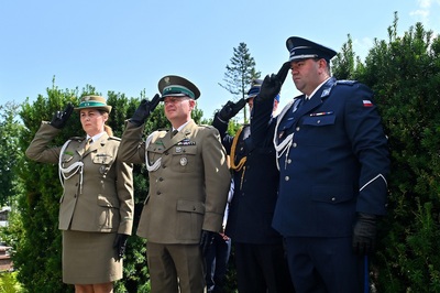 Na tle zielonych krzewów stoją w szeregu cztery osoby w mundurach wojskowych i policyjnych. Trzy osoby mają mundury wojskowe w kolorze oliwkowym z zielonymi otokami na czapkach, czwarta osoba ma granatowy mundur policyjny. Wszyscy stoją na baczność i wykonują salut wojskowy prawą ręką w rękawiczce. Na ramionach widoczne są biało-srebrne sznury galowe.