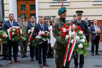 Uroczystość patriotyczna na ulicy. Na pierwszym planie żołnierz w zielonym berecie i mundurze polowym niesie duży wieniec z biało-czerwonymi kwiatami i wstęgą z napisem „Prezydent Tarnowa”. Za nim idą inni żołnierze, urzędnicy i osoby w garniturach. W tle widać osoby w mundurach i cywilów stojących w skupieniu.