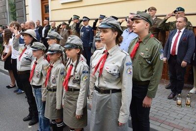 Grupa harcerzy i harcerek stoi w szeregu na ulicy. Mają szare i zielone mundury z czerwonymi chustami i naszywkami z polską flagą oraz cyfrą 5. Najmłodsze dzieci stoją na przedzie, a starsza młodzież z tyłu. Harcerze stoją na baczność, część osób w tle wykonuje salut.