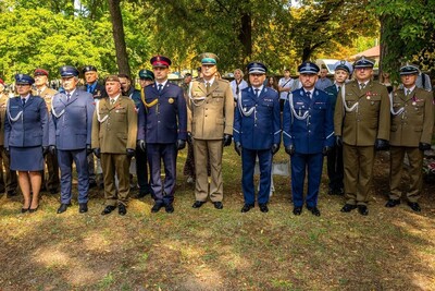 W parku na tle drzew stoi w szeregu kilkunastu oficerów w różnych mundurach wojskowych i policyjnych: oliwkowych, granatowych, niebieskich i khaki. Część ma czapki z daszkiem, inni berety. Stoją poważnie, w postawie zasadniczej, tworząc główną linię uroczystości.