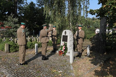 Na zdjęciu widać pięciu mężczyzn w mundurach wojskowych koloru khaki z zielonymi otokami na czapkach. Dwóch z nich stoi przy pomniku – granitowym kamieniu z tablicą pamiątkową i wieńcem z biało-czerwonymi kwiatami oraz wstążkami. Trzymają broń w pozycji honorowej. Trzeci żołnierz stoi naprzeciwko nich i salutuje, a dwóch pozostałych żołnierzy stoi obok w postawie zasadniczej. W tle widoczne są groby i krzyże cmentarne.