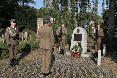 Na zdjęciu czterech żołnierzy w mundurach wojskowych stoi przy pomniku z tablicą pamiątkową i wieńcem. Dwóch żołnierzy stoi po obu stronach pomnika, trzymając karabiny. Przed pomnikiem stoją dwaj inni żołnierze – jeden z nich w postawie zasadniczej, a drugi zwrócony do pomnika, jakby oddawał cześć.