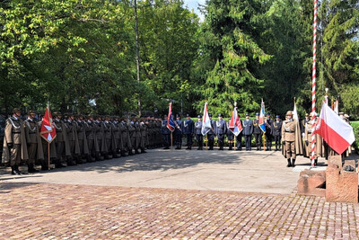 Na placu, w otoczeniu zieleni i drzew, odbywa się uroczystość wojskowa. Oddziały żołnierzy w historycznych płaszczach i rogatywkach stoją w równych szeregach. Pośrodku widoczna jest kompania reprezentacyjna z pocztami sztandarowymi – biało-czerwonymi i innymi wojskowymi flagami. Po prawej stronie stoi żołnierz przy maszcie, na którym powiewa polska flaga. Atmosfera jest podniosła i uroczysta, a ustawienie formacji wskazuje na ceremonię patriotyczną lub rocznicową.