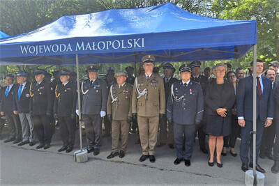 Pod dużym niebieskim namiotem z napisem &quot;Wojewoda Małopolski&quot; zgromadzili się wojskowi i przedstawiciele władz cywilnych. W pierwszym rzędzie stoją oficerowie Wojska Polskiego, Policji i innych formacji mundurowych, część z odznaczeniami i sznurami galowymi. Obok nich widoczne są osoby cywilne w eleganckich strojach. Wszyscy stoją na baczność, co sugeruje moment odśpiewania hymnu lub oddania honorów.