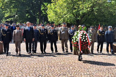 Delegacja wojskowych i urzędników w eleganckich mundurach oraz garniturach uczestniczy w ceremonii składania wieńca. Na przedzie idzie żołnierz niosący wielki wieniec ułożony z biało-czerwonych kwiatów, ozdobiony szarfą w barwach narodowych. Za nim podąża grupa oficerów i przedstawicieli władz. W tle widoczne są oddziały żołnierzy stojących w szyku, co podkreśla rangę uroczystości.