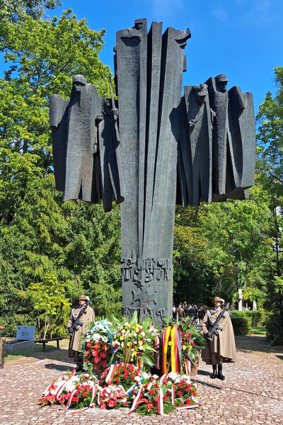 Monumentalny pomnik poświęcony Armii Krajowej, w formie wysokiej, pionowej rzeźby, przedstawiającej stylizowane postacie żołnierzy z bronią. U podstawy monumentu zgromadzone są liczne wieńce i wiązanki kwiatów w barwach narodowych Polski oraz wstęgach innych krajów, co sugeruje obecność delegacji zagranicznych. Przy pomniku stoją żołnierze w historycznych płaszczach i rogatywkach, pełniący wartę honorową. Na pomniku widnieje napis:
&quot;ARMIA KRAJOWA&quot;
