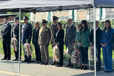 Pod dużym namiotem stoi grupa osób w dwóch rzędach. Wśród nich widać wojskowych i policjantów w galowych mundurach oraz kobiety w garniturach i garsonkach. Kilka osób trzyma owinięte w celofan wieńce z biało-czerwonymi wstęgami. Wszyscy stoją poważnie i uroczyście.