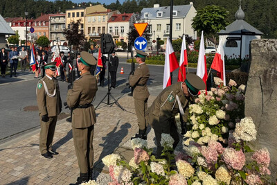 Na placu miejskim odbywa się uroczystość patriotyczna. Trzech żołnierzy w oliwkowych mundurach i zielonych czapkach stoi przy pomniku otoczonym kwiatami i biało-czerwonymi flagami. Jeden żołnierz pochyla się, aby ułożyć wieniec z kwiatami wśród białych hortensji. W tle widać ludzi z flagami i sztandarami oraz zabudowania małego rynku.