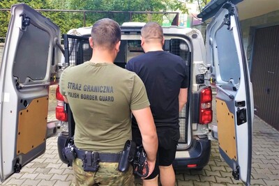 Na zdjęciu widać funkcjonariusza Straży Granicznej w zielonej koszulce z napisem „STRAŻ GRANICZNA / POLISH BORDER GUARD” na plecach. Stoi on za mężczyzną w czarnym T-shircie i czarnych spodenkach, którego prowadzi w kierunku otwartego samochodu służbowego typu więźniarka. Samochód ma otwarte tylne drzwi, wewnątrz widać metalową kratę zabezpieczającą. Funkcjonariusz ma przy pasie przypięty sprzęt służbowy, m.in. broń i kajdanki.