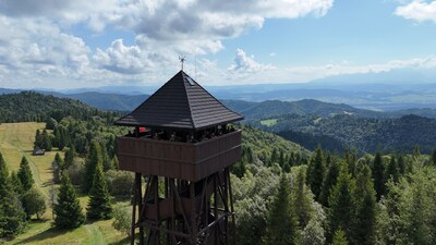 Zdjęcie panoramiczne zrobione z drona – na pierwszym planie wysoka, drewniana wieża widokowa o ciemnym dachu, pełna osób w mundurach obserwujących krajobraz. W tle piękna panorama górska z zielonymi lasami i polanami, aż po odległe, błękitne szczyty na horyzoncie.