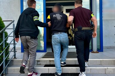 Na zdjęciu trzech mężczyzn wchodzących po schodach budynku, prawdopodobnie są to pomieszczenia biurowe placówki straży granicznej. Dwóch z nich posiada na ramionach opaski z napisem straż graniczna, prowadzą trzeciego mężczyznę, który wygląda na osobę zatrzymaną.