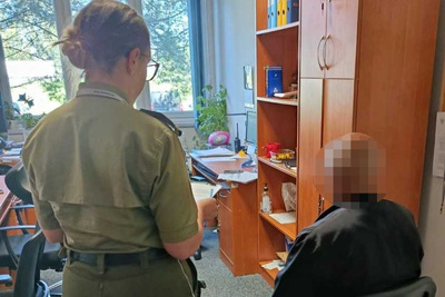 Na zdjęciu kobieta w umundurowaniu Straży Granicznej trzyma w rękach dokument. Po prawej stronie kobiety siedzący na krześle mężczyzna, prawdopodobnie osoba zatrzymana. W tle widoczne pomieszczenie biurowe z meblami biurowymi i biurkiem.