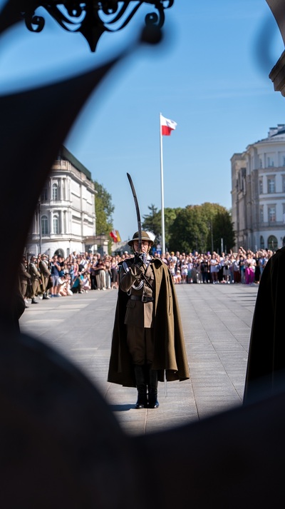 Żołnierz w tym samym podhalańskim mundurze, pelerynie i kapeluszu stoi w idealnie wyprostowanej postawie na środku placu Piłsudskiego, naprzeciw Grobu Nieznanego Żołnierza. Trzyma szablę uniesioną pionowo przed sobą, w geście oddawania honorów. W tle powiewa na masztach polska flaga narodowa, a po bokach placu widać tłum ludzi obserwujących uroczystość.
