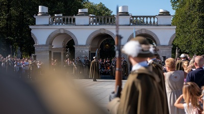 Szerokie ujęcie całego Grobu Nieznanego Żołnierza w Warszawie. Pod arkadami stoją dwaj wartownicy w mundurach, po lewej stronie widoczny jest trzeci żołnierz pilnujący porządku wśród ludzi. Po obu stronach alejki stoi tłum turystów, tworząc naturalny korytarz prowadzący wprost do grobu. Scena odbywa się w słoneczny dzień.